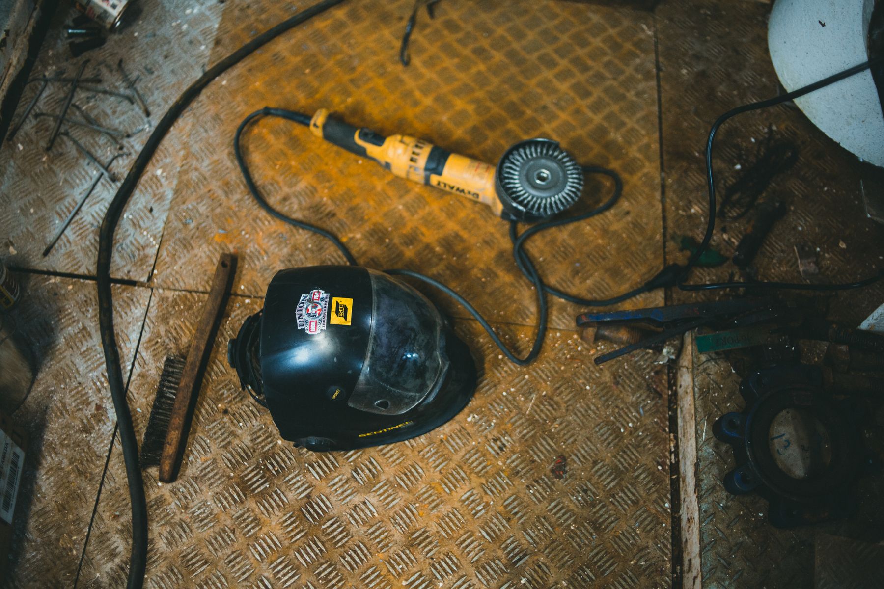 Hitsausmaski, kulmahiomakone ja teräsharja pajan kuvioidulla peltilattialla.